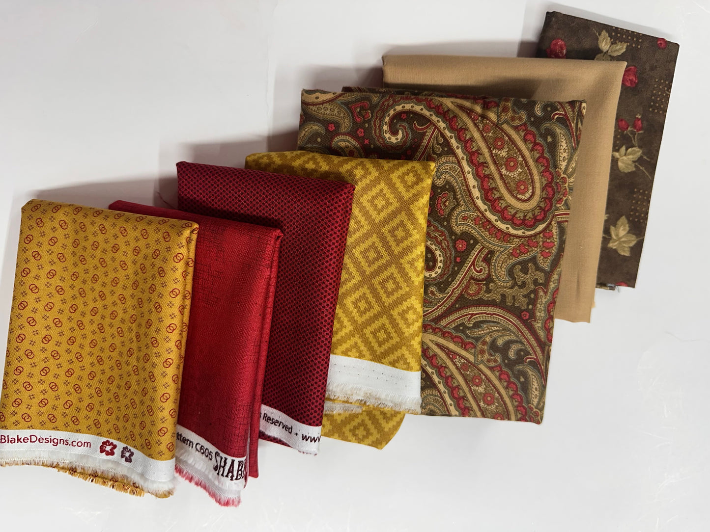 Six folded fabric swatches in various patterns and colors, including mustard yellow, deep red, gold paisley, and brown floral designs, arranged diagonally on a white background.
