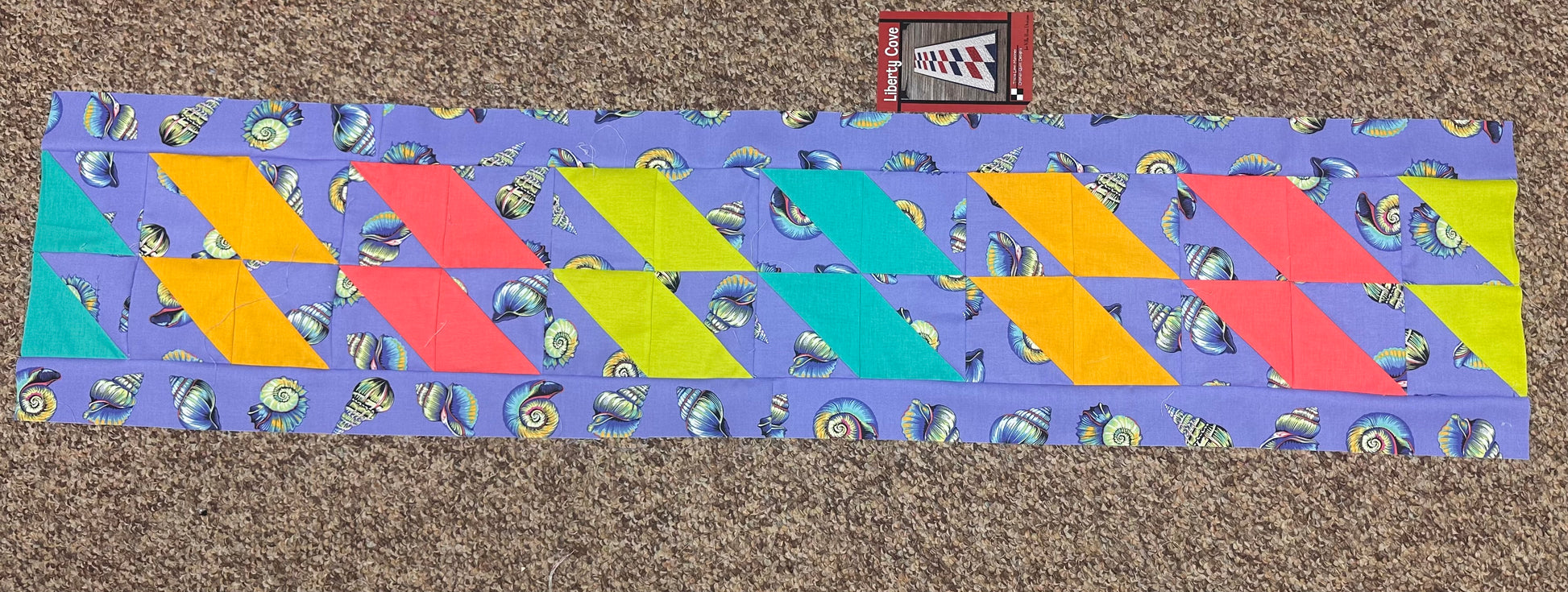 A colorful geometric pattern of pink, yellow, teal, and orange diamond shapes on a lavenderLil Shelly coral reef  background.