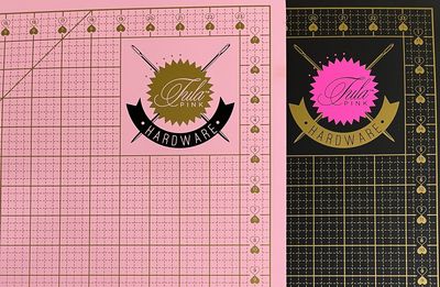 A double sided cutting mat with one side pink and the other black, both with gold grid markings, and the Tula Pink brand logo on both sides.